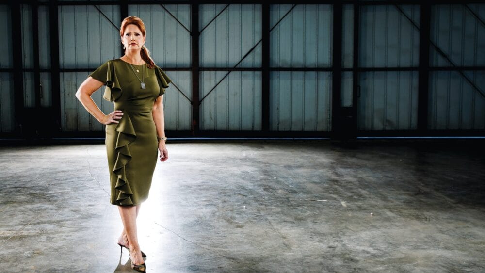 A woman in a green, ruffled dress and heels stands confidently in a large, empty, industrial space with sunlight streaming in from behind her. South Florida Business & Wealth