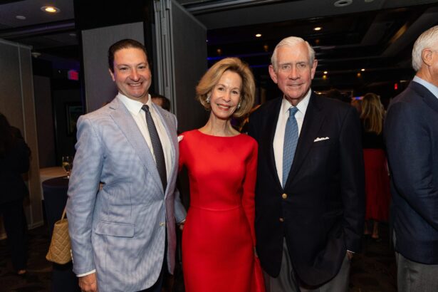 Three adults pose together at an indoor event; the man on the left wears a light blue suit, the woman in the center wears a red dress, and the man on the right wears a dark suit and light blue tie. South Florida Business & Wealth