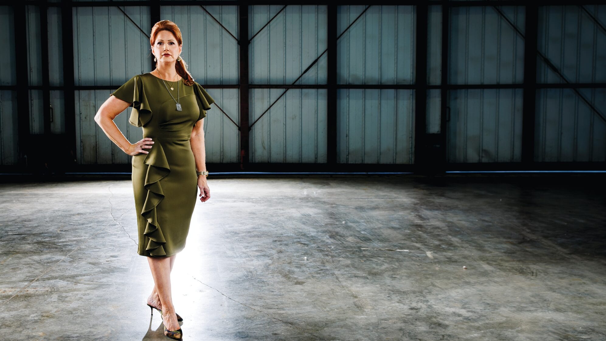 A woman in a green, ruffled dress and heels stands confidently in a large, empty, industrial space with sunlight streaming in from behind her. South Florida Business & Wealth