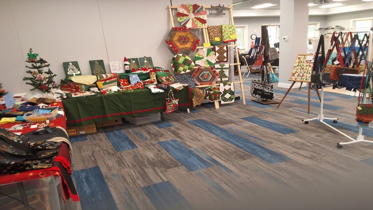 A room displays handmade crafts, including quilted items, fabric bags, table runners, and small decorated Christmas trees. The crafts are arranged on tables and racks in a well-lit space with blue and gray flooring. South Florida Business & Wealth