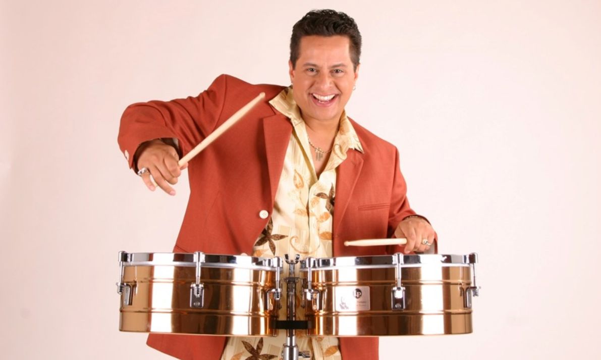 A smiling man in a red blazer and patterned shirt plays a pair of gold timbales with drumsticks against a plain light background. South Florida Business & Wealth