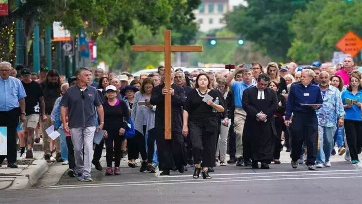 A large group of people walks down a city street during the day, with several individuals at the front carrying a large wooden cross. Many are dressed in dark clothing, and some wear clerical attire. Trees line the street in the background. South Florida Business & Wealth