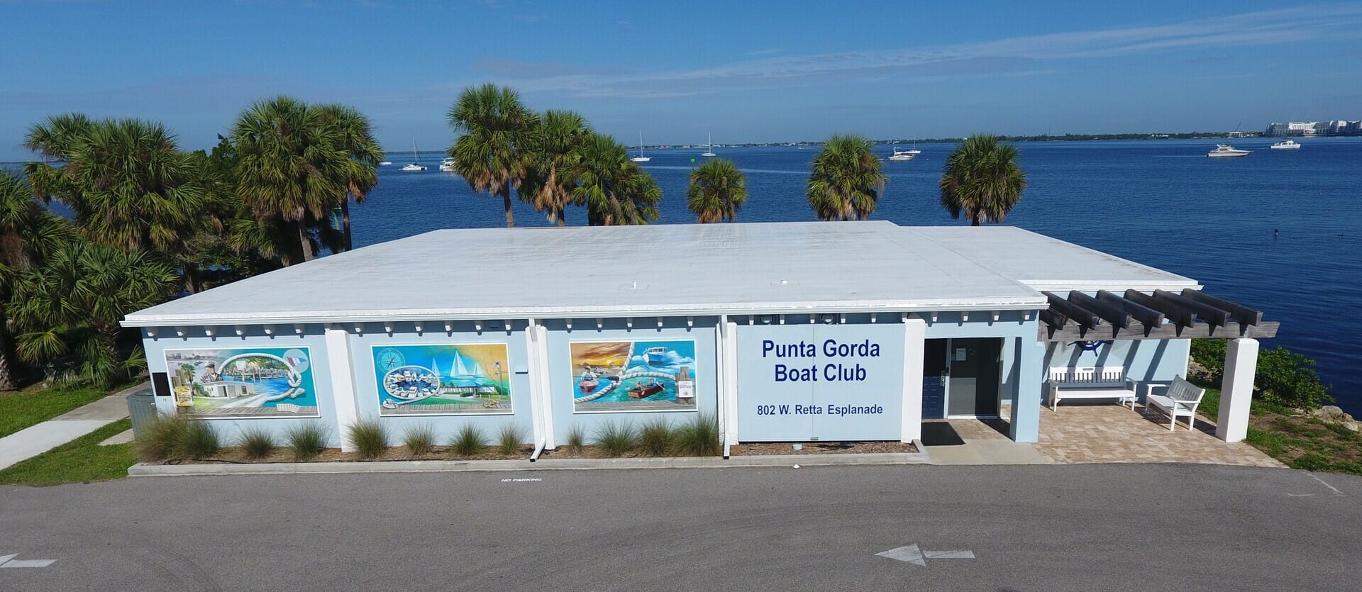 A white building labeled "Punta Gorda Boat Club" sits by the water, with palm trees nearby, colorful murals on its side, and several boats on the blue bay in the background under a clear sky. South Florida Business & Wealth
