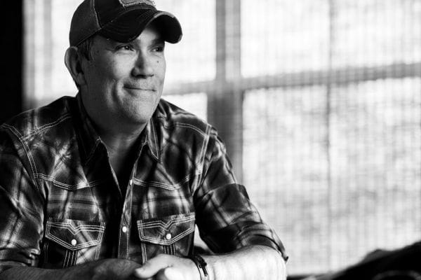 Black and white photo of a smiling man wearing a plaid shirt and a baseball cap, sitting indoors with his arms resting on a table. Sunlight filters through a window in the background. South Florida Business & Wealth