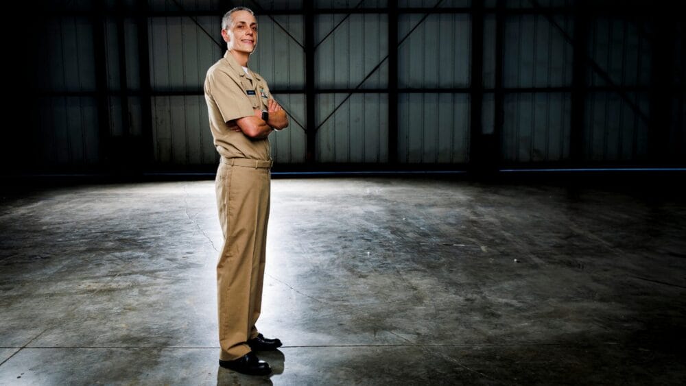 A person in a tan military uniform stands with arms crossed in a large, empty industrial space with a concrete floor and a dark, metal-paneled wall in the background. South Florida Business & Wealth