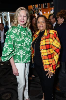 Two women stand side by side at an indoor event. One wears a green and white patterned blouse with cream pants, and the other wears a bright orange and yellow plaid jacket over a black outfit. People mingle in the background. South Florida Business & Wealth