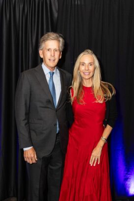 A man in a dark suit and blue tie stands next to a woman in a bright red dress and black cardigan. They are smiling and posing in front of a black curtain backdrop. South Florida Business & Wealth