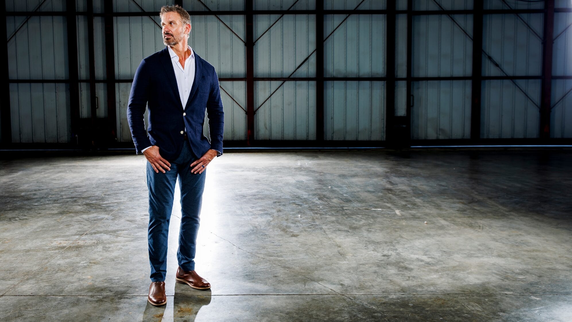 A man in a navy blazer, white shirt, and blue pants stands on a polished concrete floor inside a large, empty industrial warehouse with metal walls and minimal lighting. South Florida Business & Wealth
