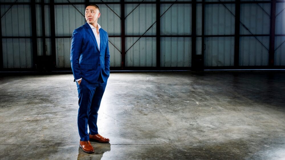 A man in a blue suit and brown shoes stands with hands in pockets on a polished concrete floor in a spacious, industrial warehouse, looking to his left. South Florida Business & Wealth