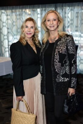 Two women, both with long blonde hair, stand side by side indoors. One wears a black jacket and pleated skirt, holding a beige handbag; the other wears a black top with a decorative black and silver jacket, holding a black purse. South Florida Business & Wealth