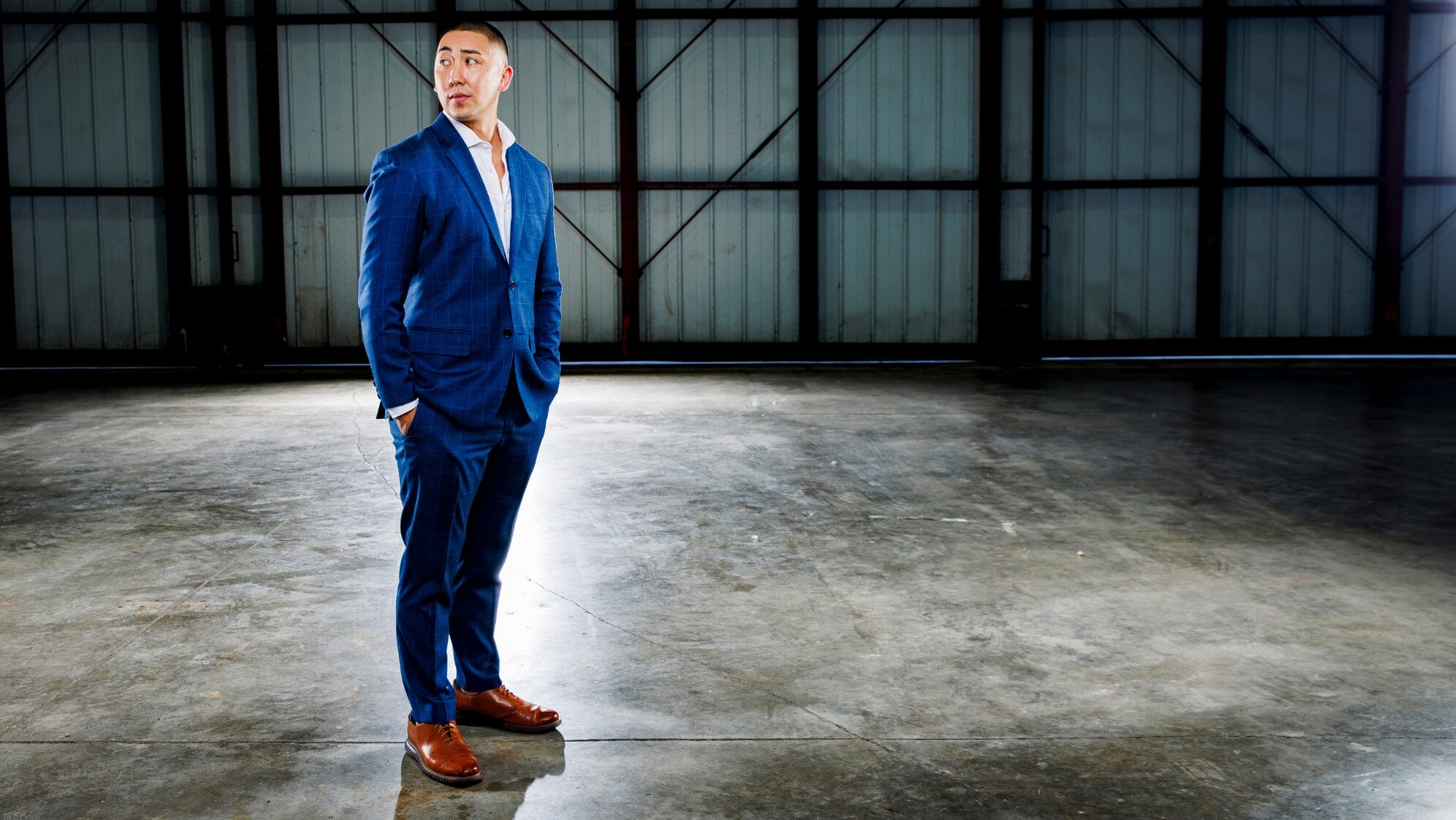 A man in a blue suit and brown shoes stands with hands in pockets on a polished concrete floor in a spacious, industrial warehouse, looking to his left. South Florida Business & Wealth