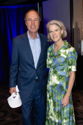 A man in a blue suit and a woman in a green and blue floral dress stand together indoors, smiling at the camera. The man holds a white hat in his hand. The background is dimly lit with blue and dark tones. South Florida Business & Wealth