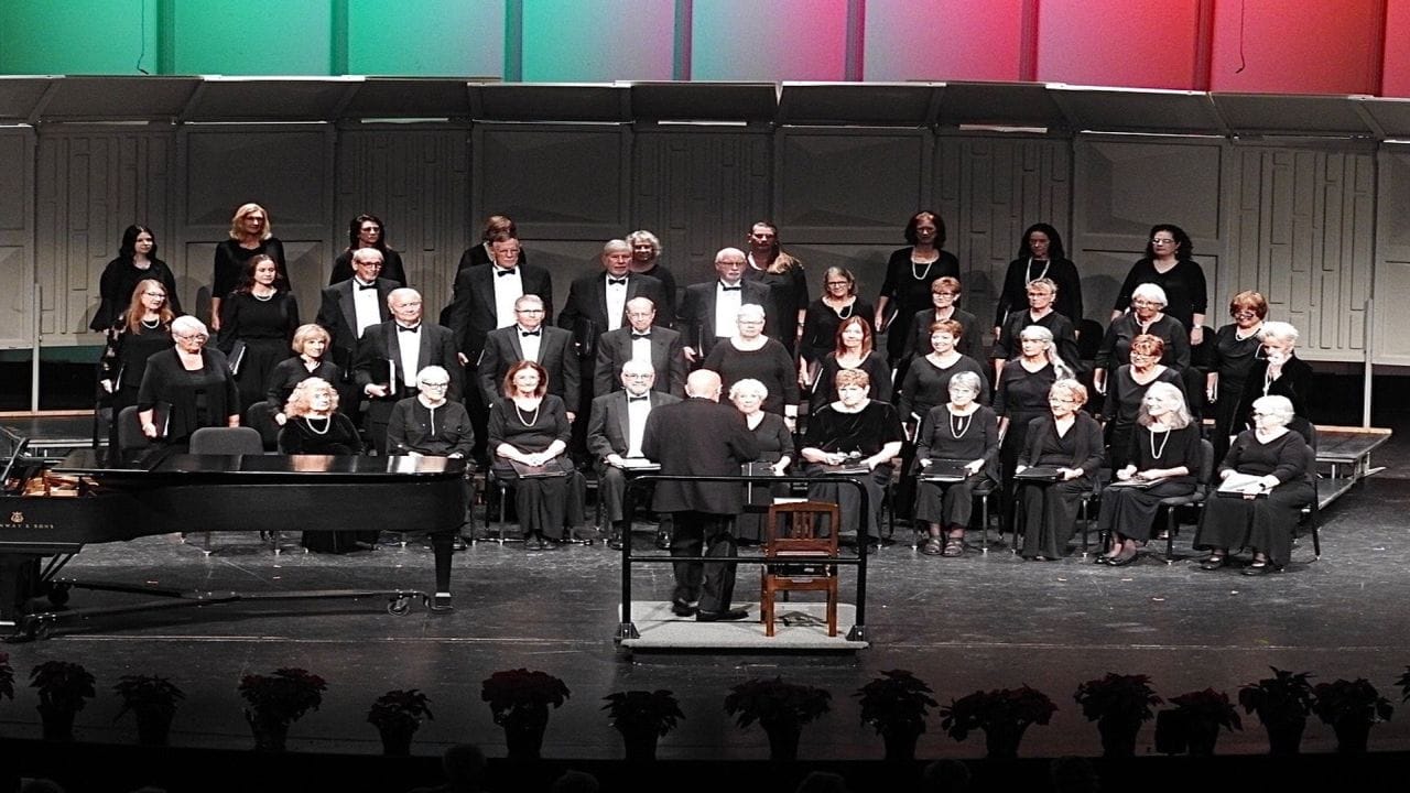 A choir of men and women dressed in black performs on a stage under red, white, and green lights, with a conductor in front and a grand piano to the side. The background has tiered risers and a white acoustic shell. South Florida Business & Wealth
