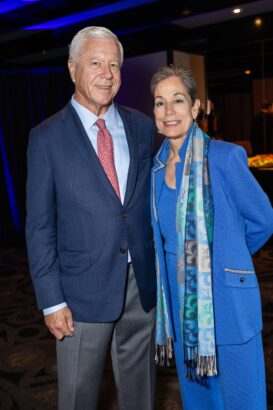 An older man in a blue suit and red tie stands next to an older woman in a blue outfit and patterned scarf. They are smiling and posing together indoors at a formal event. South Florida Business & Wealth
