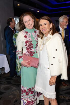 Two women stand together and smile at an indoor event. One wears a long floral dress and holds a pink clutch, while the other wears a white coat with pearls. Other people are mingling in the background. South Florida Business & Wealth