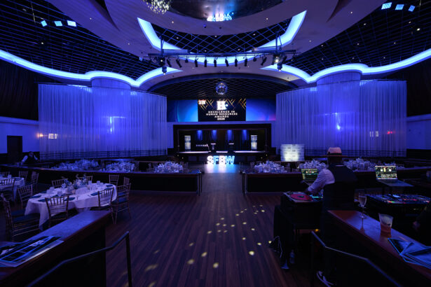 A spacious, elegant event venue is set up for an awards show, with round dinner tables, blue accent lighting, sheer white curtains, and a stage displaying "Excellence Awards 2023" in front of a large screen. South Florida Business & Wealth