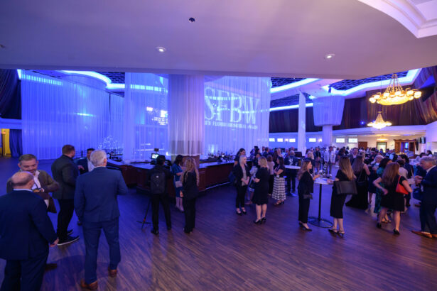 A large group of people in formal attire mingle at a spacious, elegant event venue with blue lighting, sheer curtains, and a chandelier. A bar and a projected logo are visible in the background. South Florida Business & Wealth