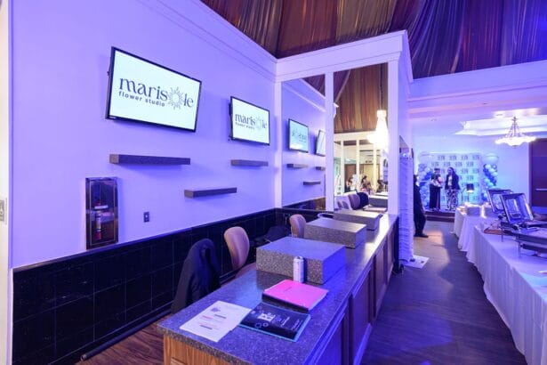 Reception area with granite counters, clipboards, and a can; three wall-mounted screens display "marisole flower studio." In the background, people gather near event tables and balloon decorations under elegant lighting. South Florida Business & Wealth