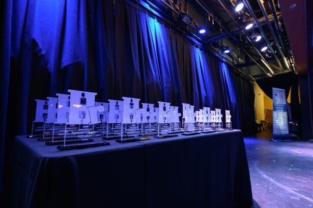 A row of clear acrylic awards is arranged on a black table, illuminated by blue stage lighting, with dark curtains and stage equipment visible in the background. South Florida Business & Wealth