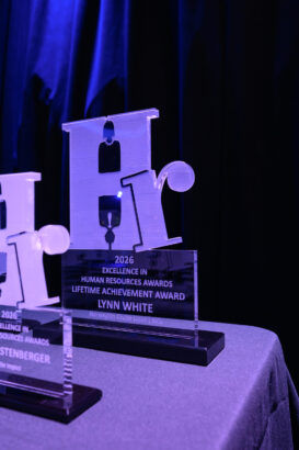 A glass award trophy engraved with "2026 Excellence in Human Resources Awards Lifetime Achievement Award Lynn White" sits on a table against a dark background. Another similar trophy is partially visible beside it. South Florida Business & Wealth