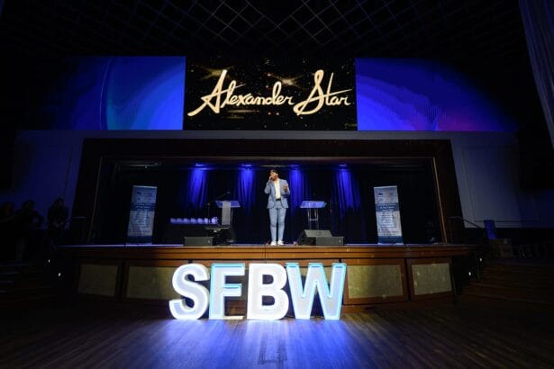 A person in a light blue suit stands on stage under a large screen displaying "Alexander Star," with illuminated "SFBW" letters on the floor and blue lights highlighting the background. South Florida Business & Wealth