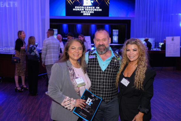 Three smiling people pose together at an awards event. The woman on the left holds a plaque. Others are mingling in the background. A screen reads “Excellence in Human Resources Awards 2024.”. South Florida Business & Wealth