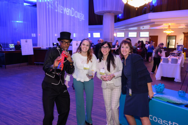 Four people pose and smile at an indoor event; one is dressed as a musician in a top hat playing a saxophone, while the others hold drinks and stand by a blue table with informational materials. The atmosphere is festive and casual. South Florida Business & Wealth