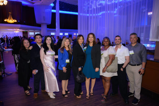 A group of nine people dressed in business or cocktail attire pose together and smile for a photo at an indoor event with blue lighting and other attendees in the background. South Florida Business & Wealth