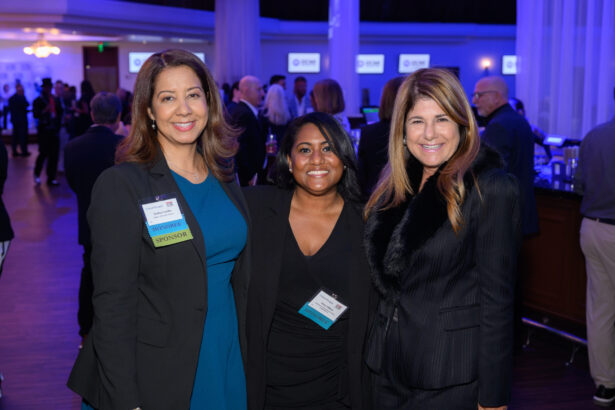 Three women in professional attire smile at the camera during a networking event; people and screens are visible in the blue-lit background. South Florida Business & Wealth