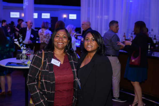 Two women in business attire smile at the camera at a networking event. Several people are socializing in the background near a bar, with blue ambient lighting and high-top tables visible. South Florida Business & Wealth