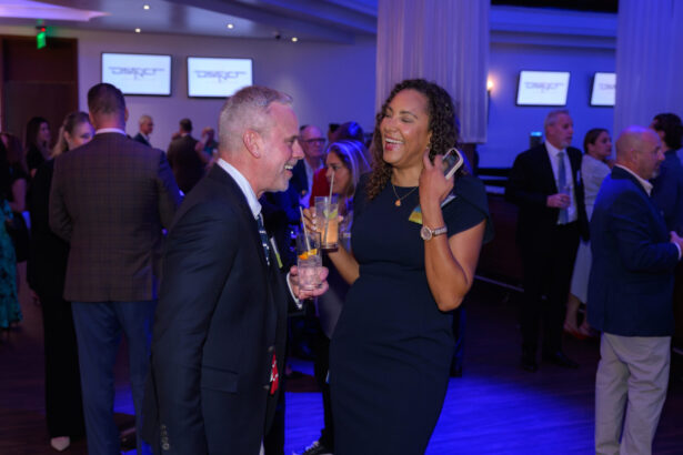 Two people dressed formally are laughing and holding drinks at a lively indoor event, with other guests socializing in the background under blue lighting. South Florida Business & Wealth
