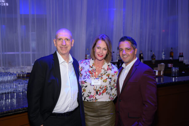 Three people dressed in business attire stand together smiling in front of a bar with bottles and glasses, with blue lighting and sheer curtains in the background. South Florida Business & Wealth