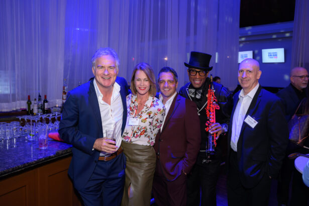 Five people pose together, smiling at an indoor event with blue lighting. They are dressed in semi-formal to formal attire. A bar with glasses and bottles is in the background, and two screens are visible on the far wall. South Florida Business & Wealth
