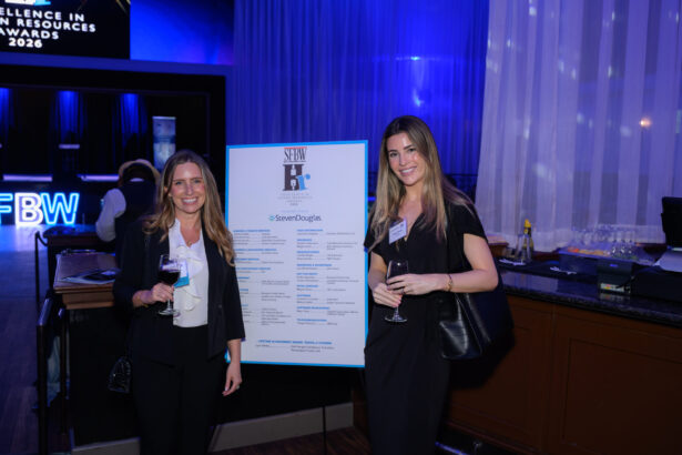 Two women dressed in business attire stand and smile beside an event sign, each holding a glass of wine, in a dimly lit venue with blue lighting and a bar in the background. South Florida Business & Wealth