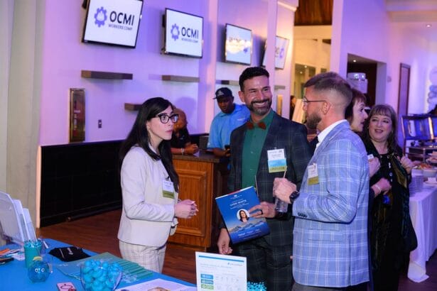 Four people in business attire converse at a conference booth with brochures and branded items. Monitors display "OCMI WORKSHOP 2023." The setting is brightly lit and modern. South Florida Business & Wealth