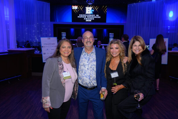 Four people dressed in business attire smile and pose for a photo at an indoor event, with drinks in hand. A sign behind them reads "Excellence in Human Resources Awards 2026." The room is lit with blue lighting. South Florida Business & Wealth