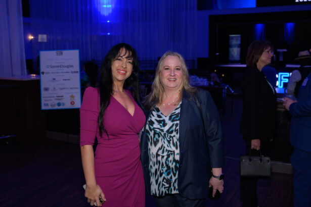 Two women pose and smile for a photo at an indoor event with blue lighting. One wears a magenta dress, the other a dark blazer over a patterned top. People and an event sign are visible in the background. South Florida Business & Wealth