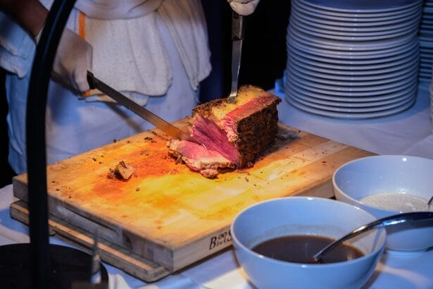 A chef in white gloves slices a large roast beef on a wooden cutting board, with two bowls of sauce and a stack of white plates nearby on a table. South Florida Business & Wealth