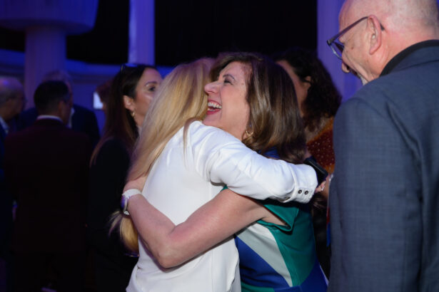 Two women happily embrace at a lively indoor event, surrounded by other people who are smiling and talking, with blue lighting creating a festive atmosphere. South Florida Business & Wealth