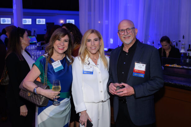 Three people dressed in business attire smile and pose together at an indoor event. A woman holds a glass of wine, and event name badges are visible. The background shows a bar area and other attendees. South Florida Business & Wealth