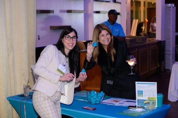 Two women smiling and holding blue promotional items while standing at an event table with informational materials. One holds a wine glass; both wear business attire. A man stands in the background near a bar area. South Florida Business & Wealth
