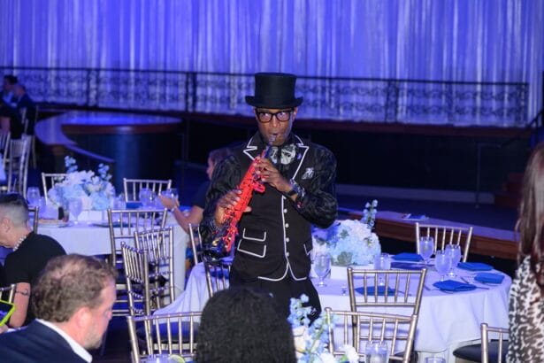 A musician dressed in a black suit and top hat plays a red saxophone while walking between tables at an elegant, formal event with white and blue decor. South Florida Business & Wealth