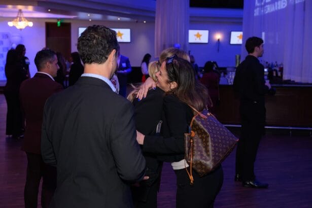 A woman with a brown designer backpack hugs another person at an indoor formal event, surrounded by people in suits and dresses. The background includes a bar, screens with star icons, and dim blue lighting. South Florida Business & Wealth