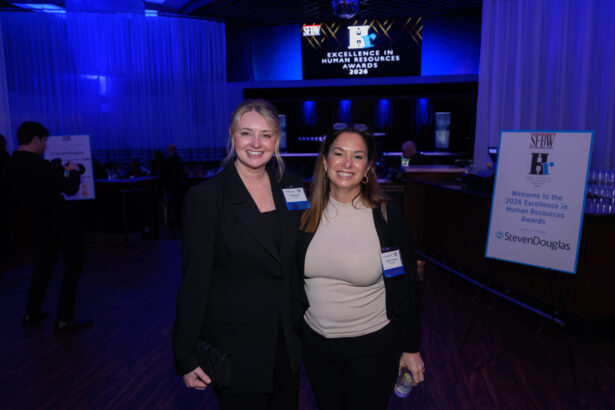Two women smile and pose together at an indoor event. Behind them, a large screen displays “Excellence in Human Resources Awards 2026.” An event sign and other attendees are visible in the background. South Florida Business & Wealth
