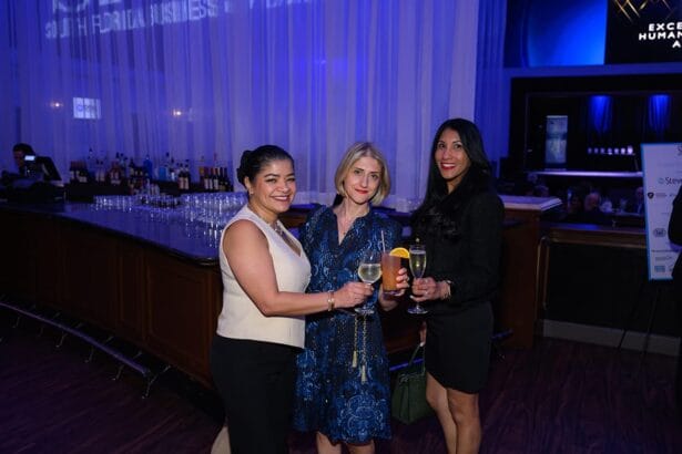 Three women dressed formally stand together holding drinks and smiling at an indoor event with a bar and a blue-lit background. South Florida Business & Wealth