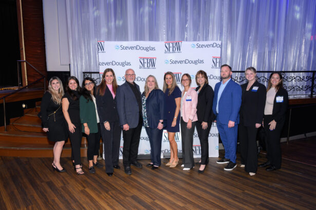 A group of twelve people, dressed in business attire, stand in a line and smile at the camera in front of a backdrop with "SFBW" and "StevenDouglas" logos at an indoor event. South Florida Business & Wealth