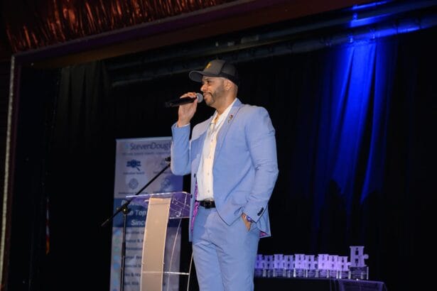 A man in a light blue suit and black cap speaks into a microphone on stage, standing next to a clear podium, with a blue-lit background and a banner behind him. South Florida Business & Wealth