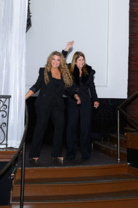 Two women wearing black suits smile and wave as they walk down a small set of stairs indoors, next to a white curtain and a railing. South Florida Business & Wealth