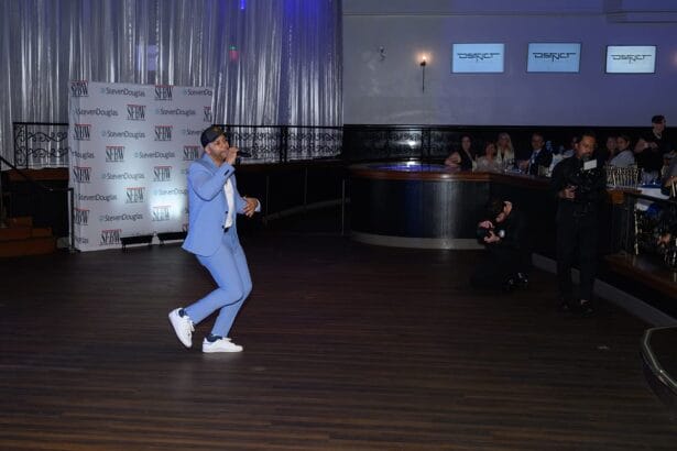 A man in a light blue suit and white sneakers performs on stage in a spacious venue with wood flooring. Photographers and audience members watch, and logos and banners are visible in the background. South Florida Business & Wealth