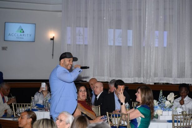 A man in a light blue jacket and black cap sings into a microphone while walking through a seated crowd at an indoor event. Guests sit at tables, some smiling and taking photos. White drapes and monitors are in the background. South Florida Business & Wealth